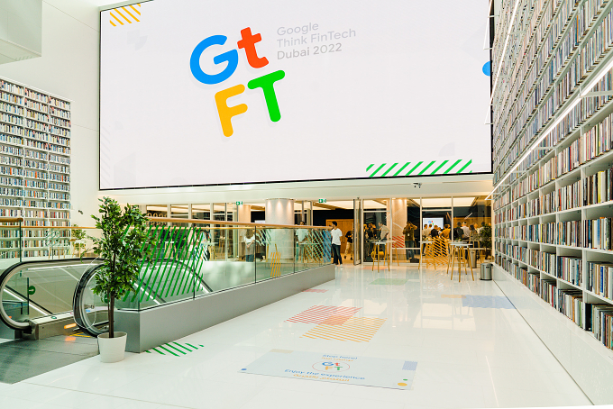 Atrium entrance with a giant on-site screen displaying the GtFT logo, flanked by bookshelves and an escalator